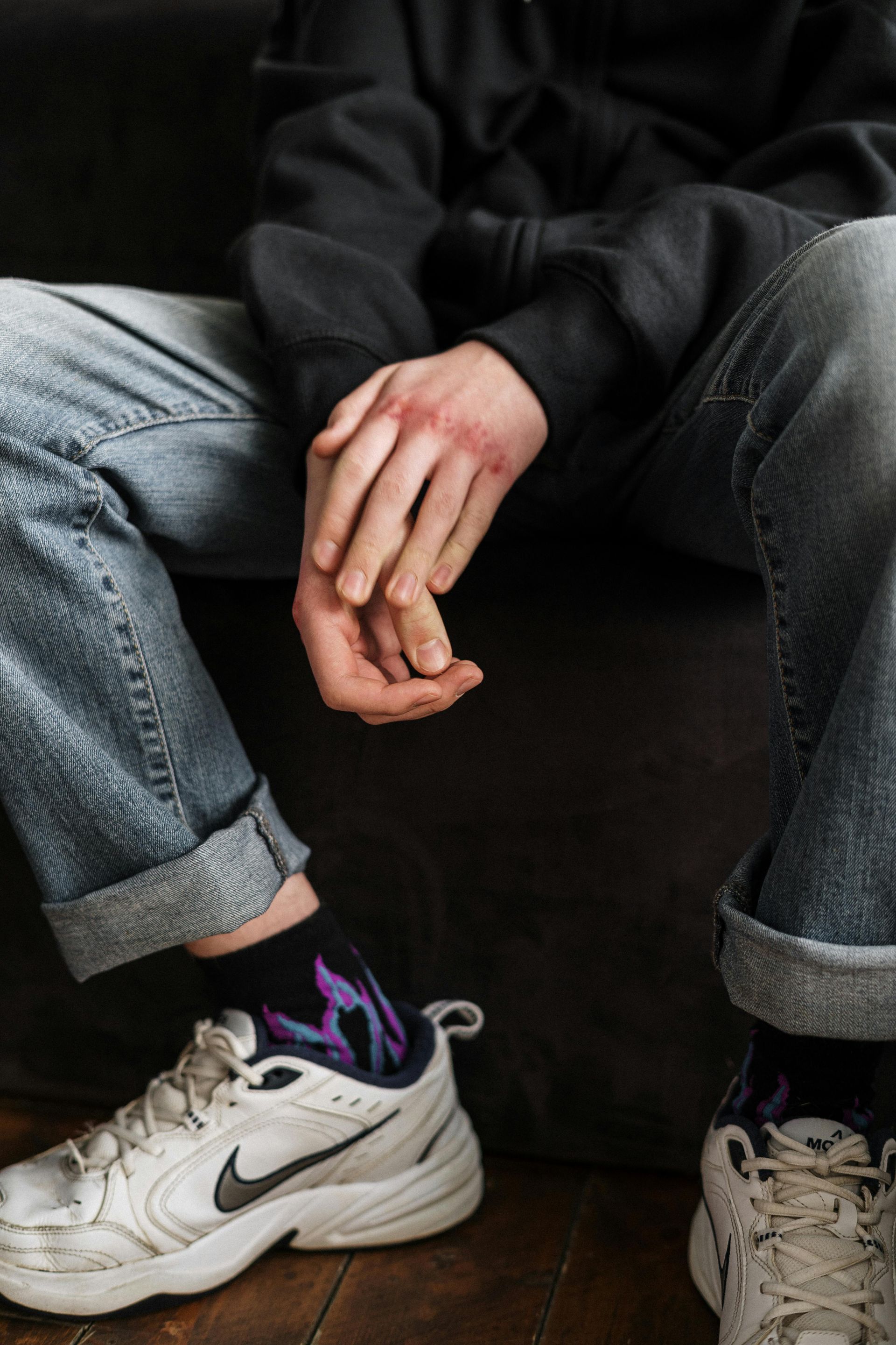 Person sitting on a couch, hands clasped, wearing cuffed jeans and white sneakers.