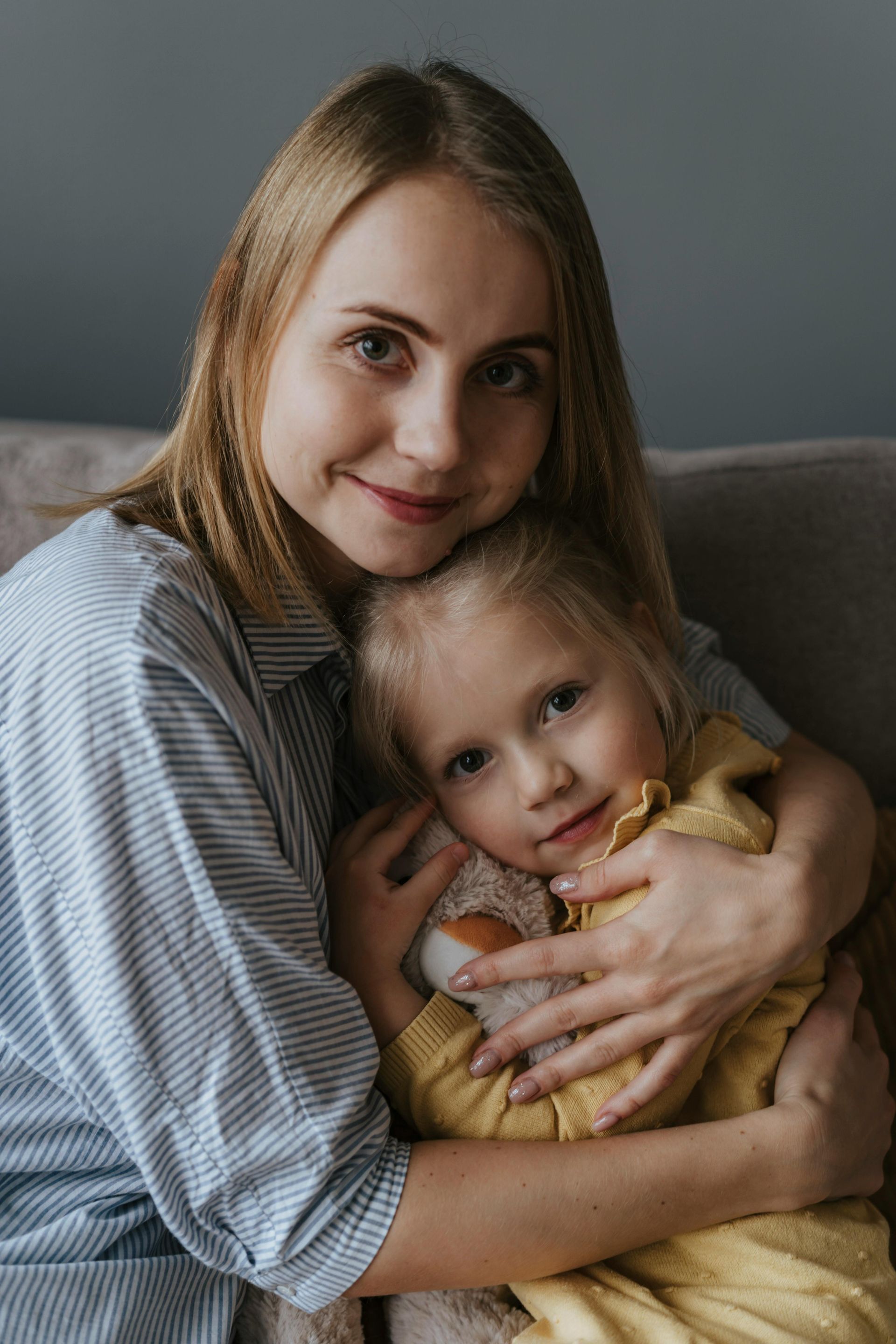 Woman hugging a small child on a couch, both looking at the camera, in soft natural light.