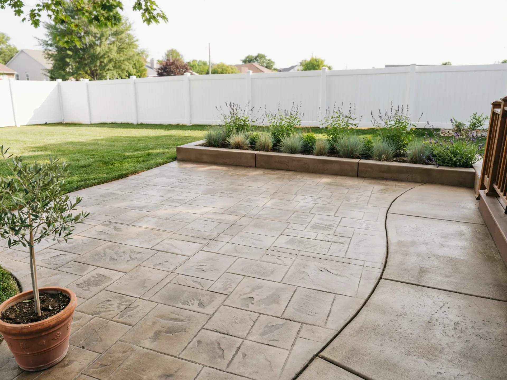 Backyard patio with stamped concrete, potted tree, raised planter, and white fence under a bright sky