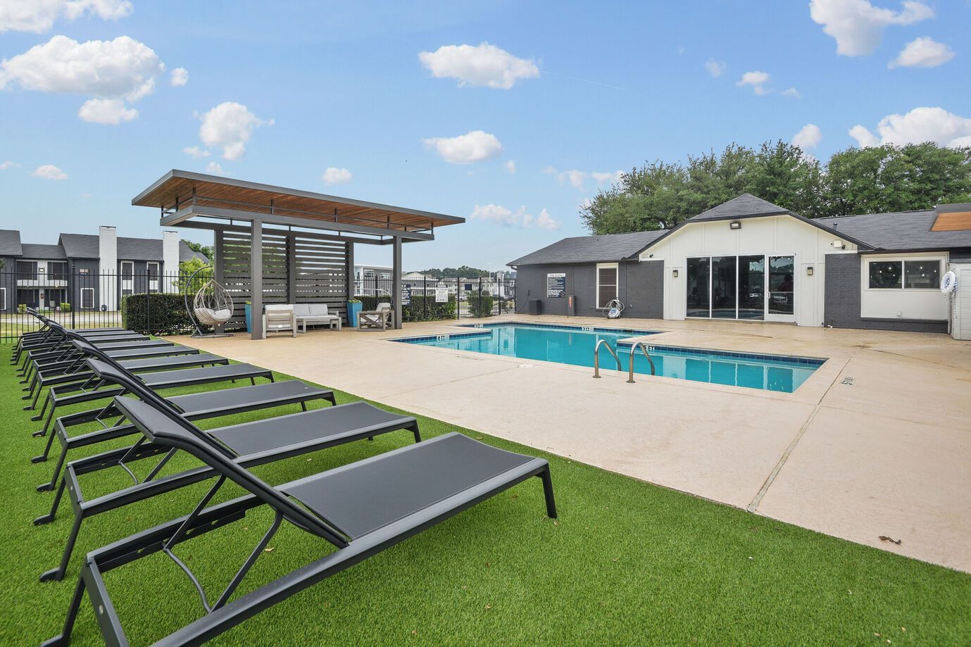 Poolside scene with lounge chairs, a pool, and a shaded seating area under a blue sky.