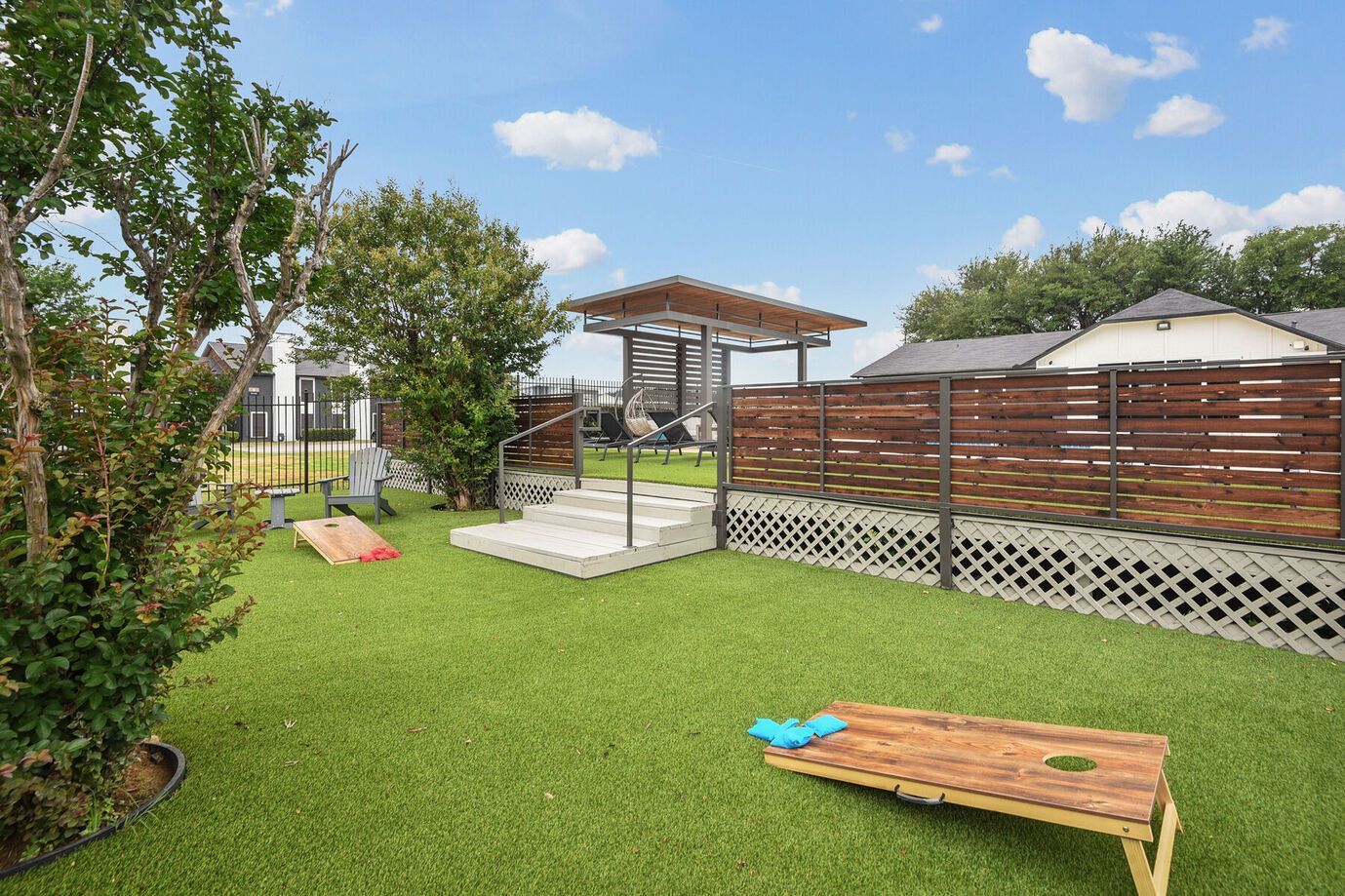 Dog park with artificial turf, wooden fence, and pergola. Cornhole game set up on the grass.
