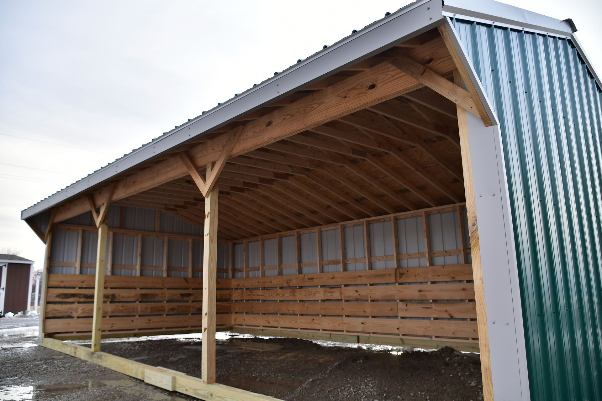 Wooden open-sided shelter with a sloped metal roof and concrete base, viewed from the front-left.