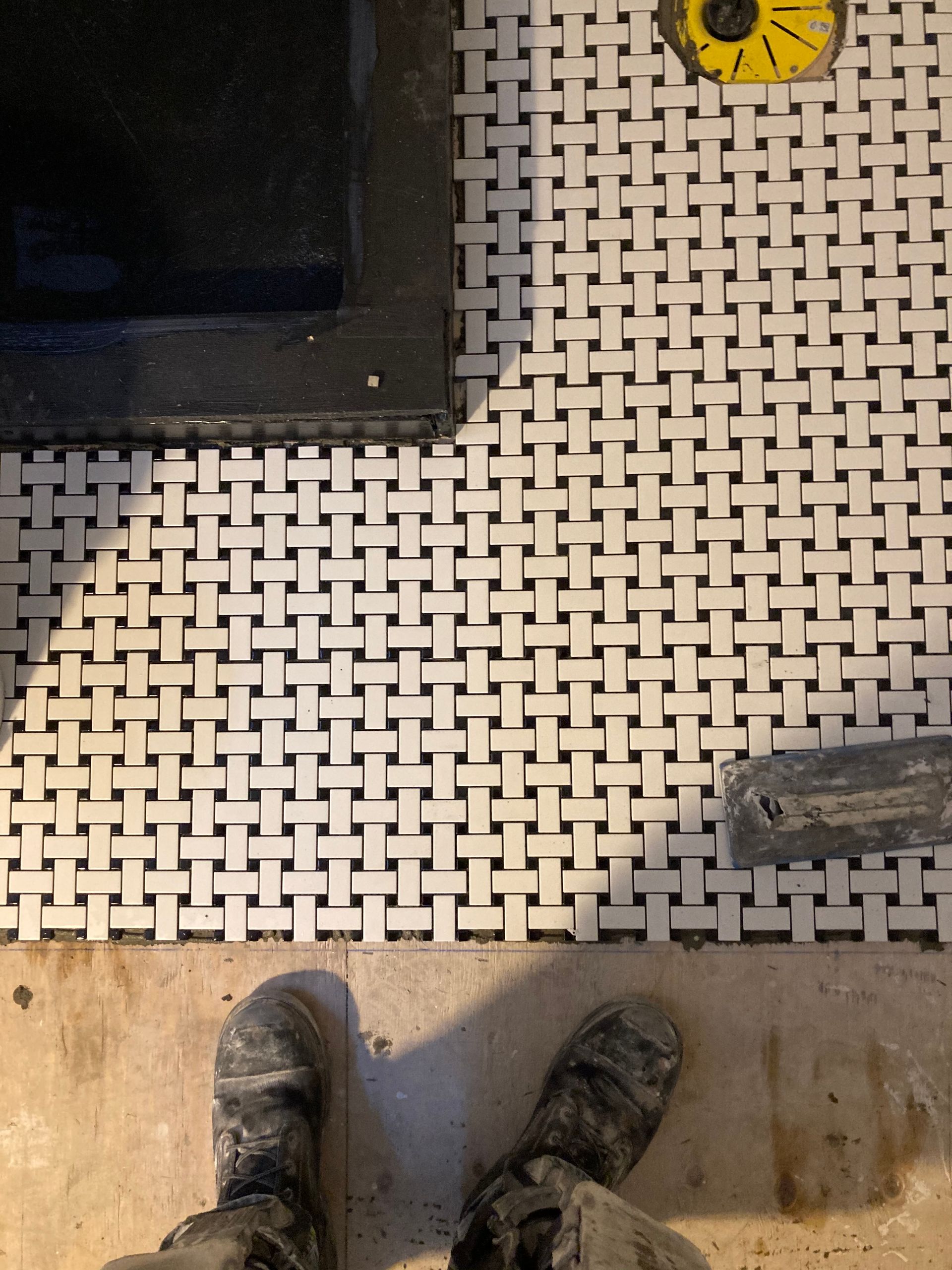 Les pieds d'une personne se tiennent debout sur un sol carrelé à motifs avec des joints noirs, près d'un objet noir.