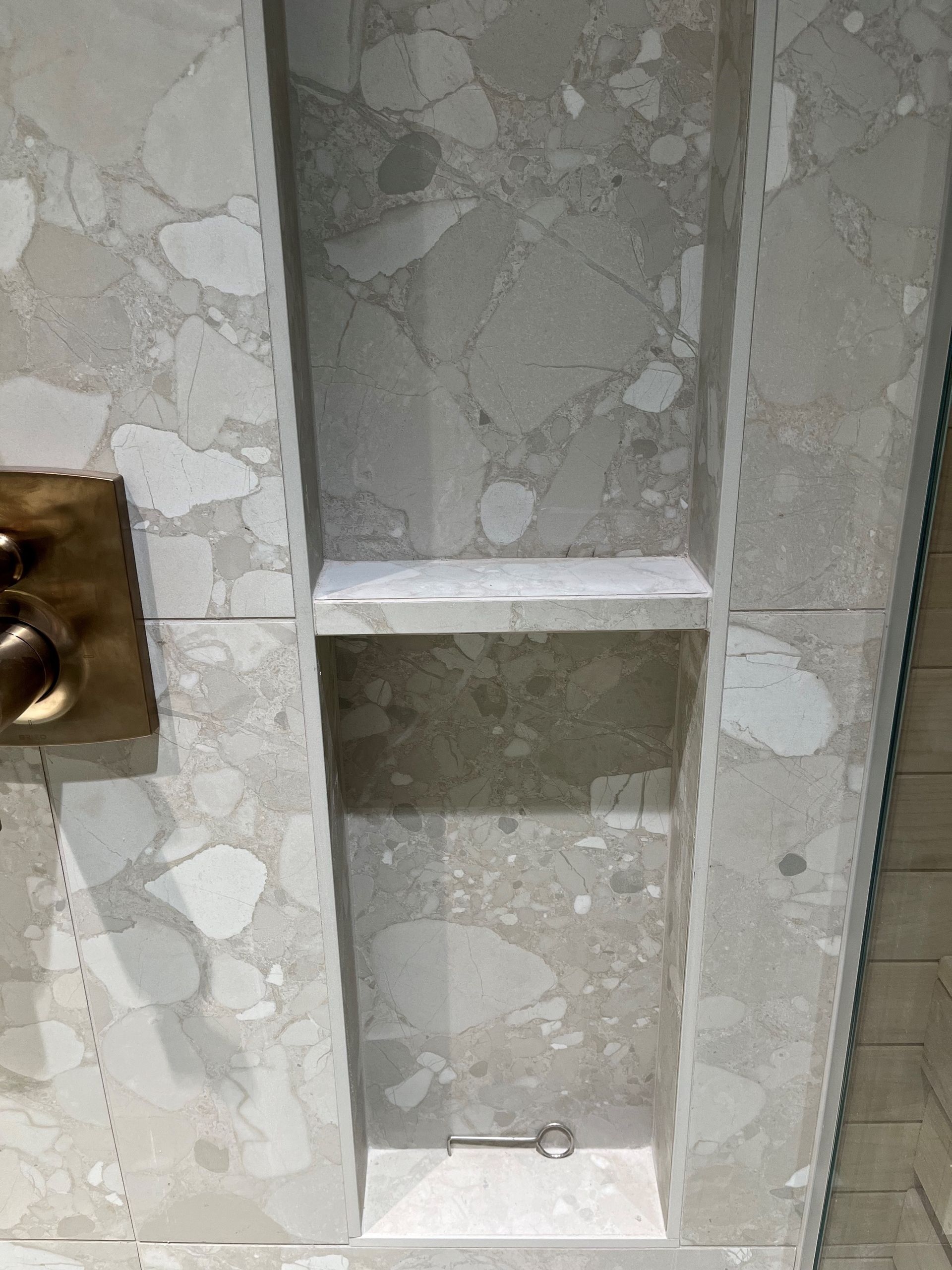 Paroi de douche avec étagère intégrée, carrelée de pierre mouchetée gris clair et blanche, robinetterie de douche dorée.