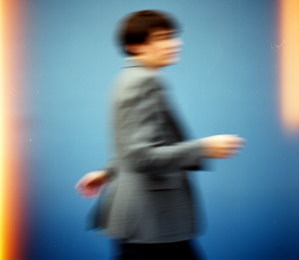 Blurred person walking past a blue wall, wearing a gray jacket
