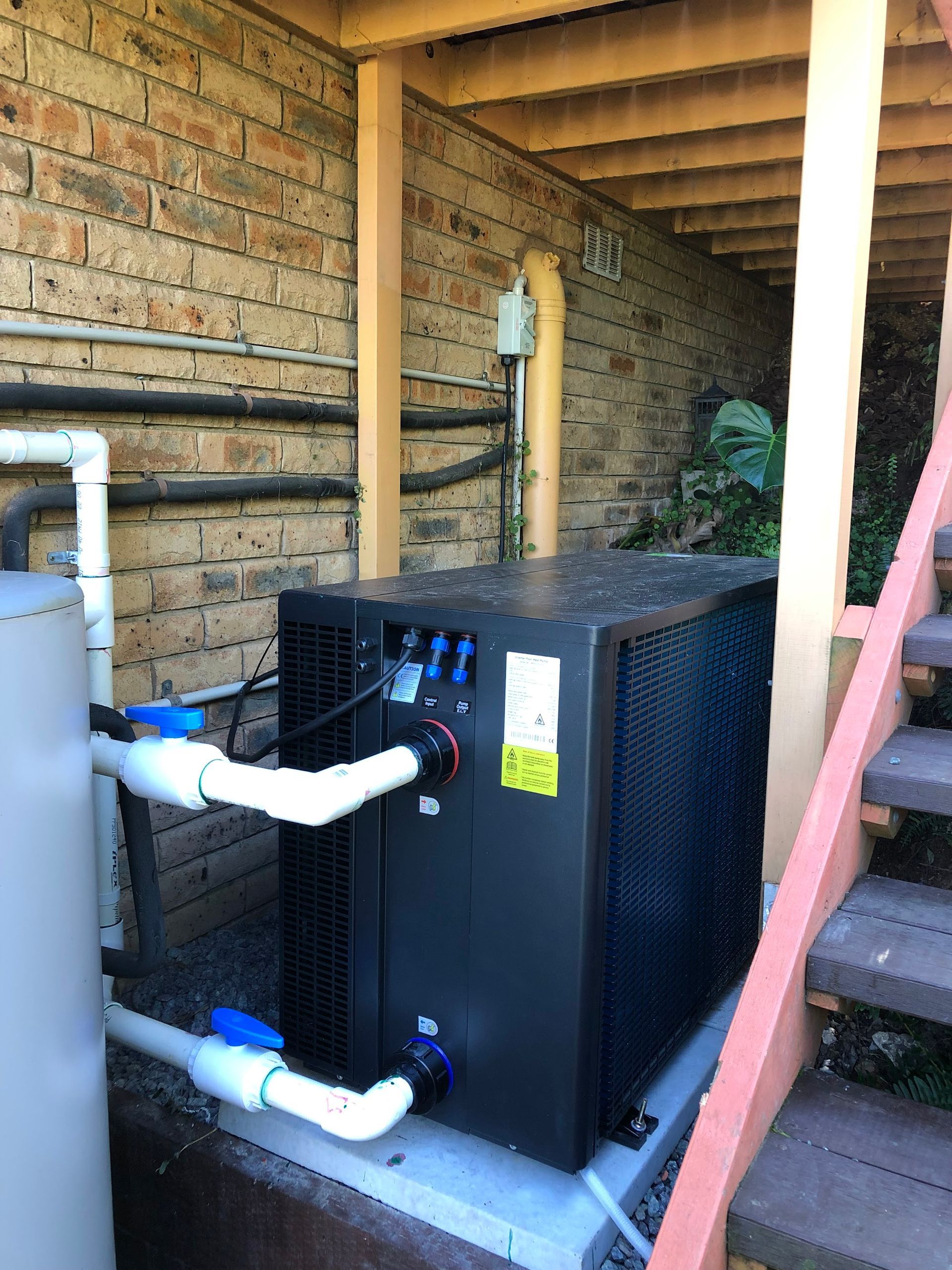 Outdoor HVAC unit beside a brick wall under a wooden porch, with white pipes and stairs nearby — Plumbing in Canberra, ACT