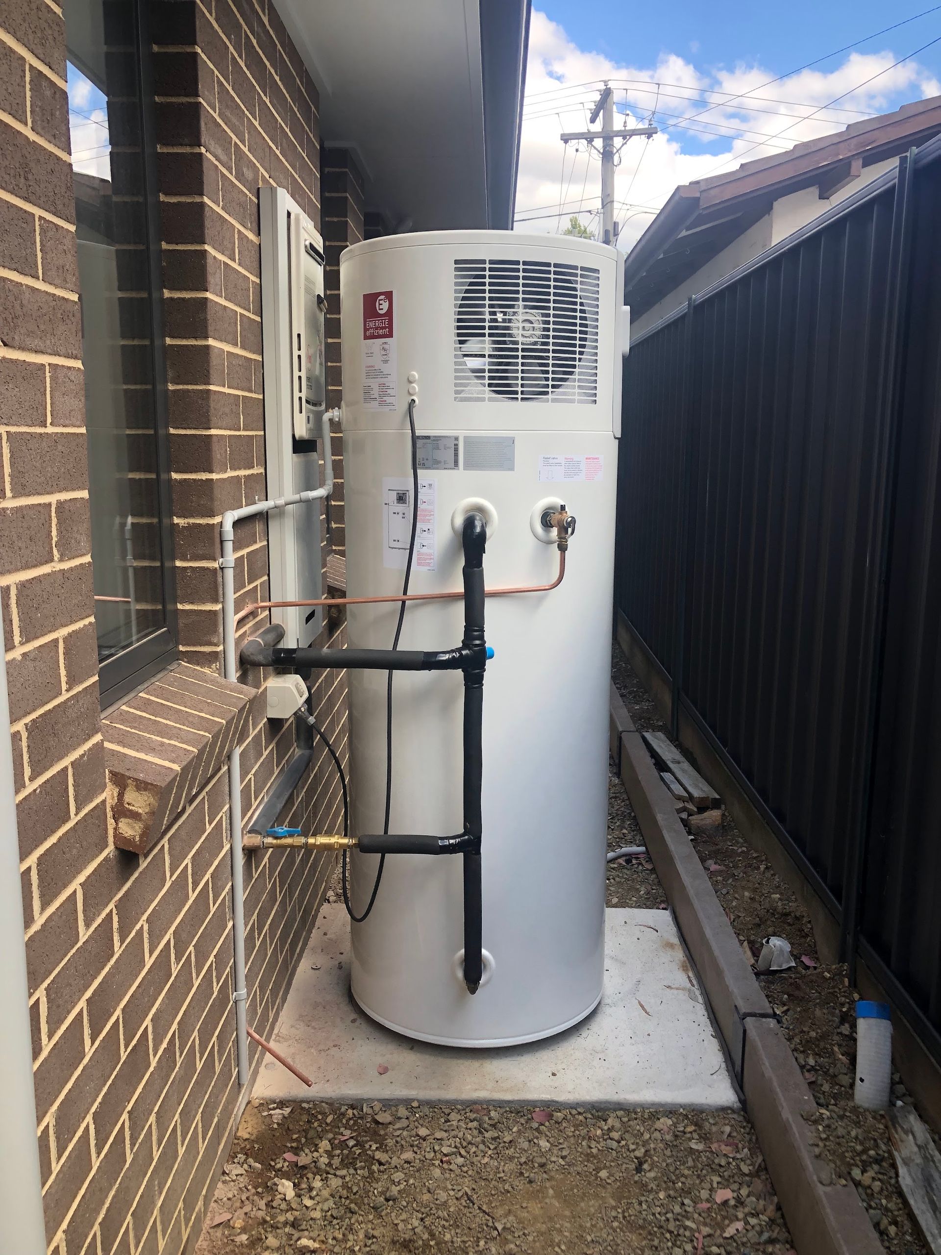 White outdoor water heater beside brick house and fence in narrow side yard — Plumbing in Canberra, ACT