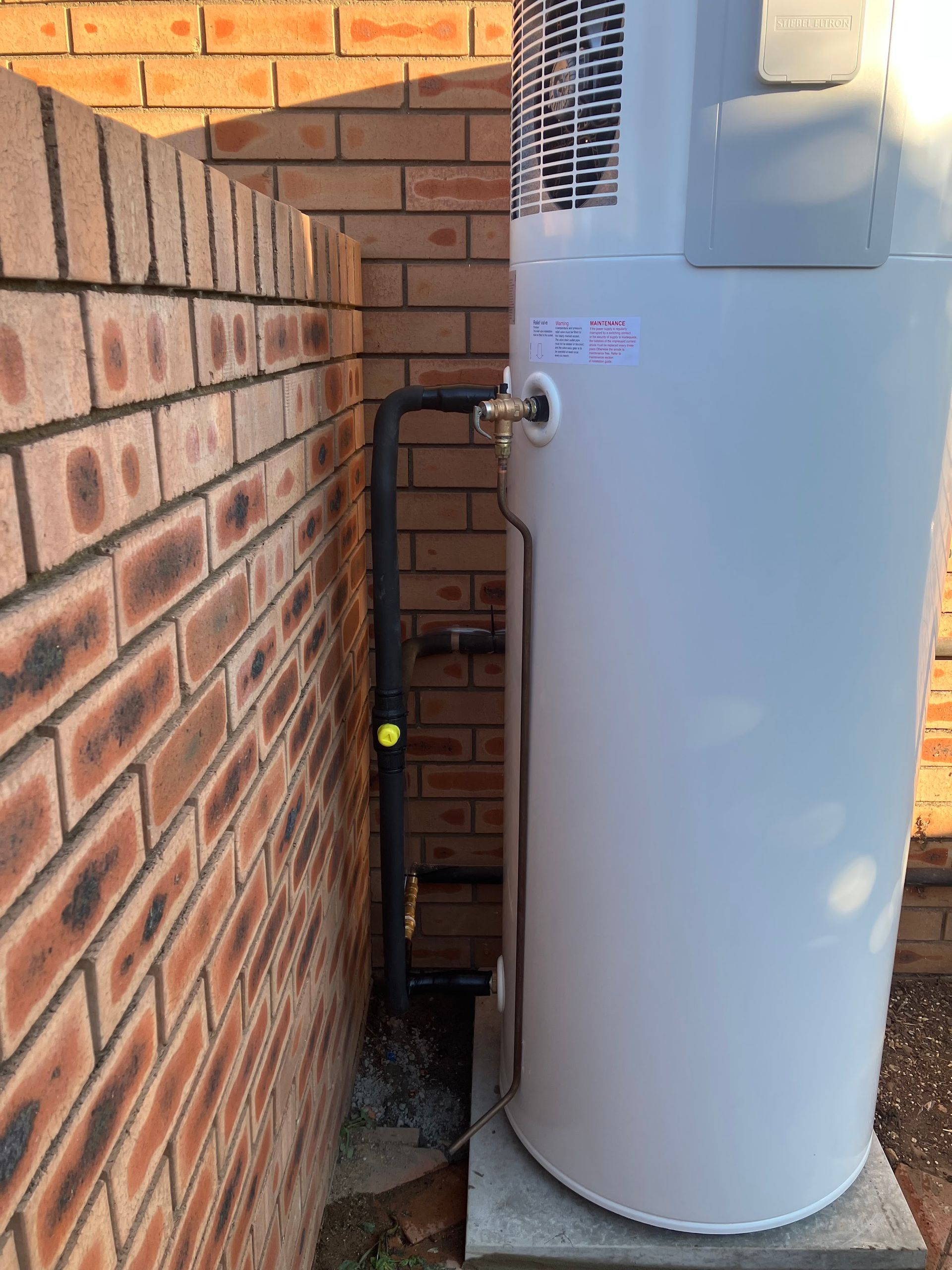 Water heater beside a brick wall in a narrow outdoor utility area, with exposed pipes and sunlight — Plumbing in Canberra, ACT
