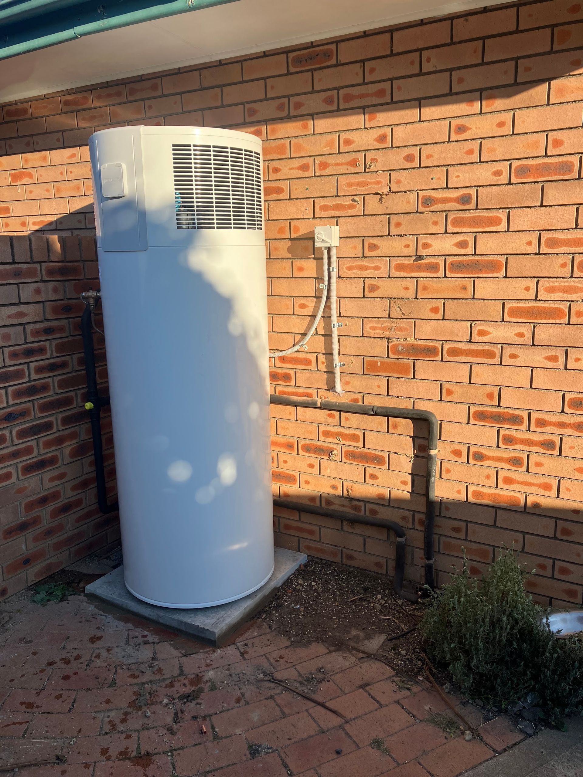 White outdoor utility unit beside a brick wall with exposed pipes in a sunlit courtyard — Plumbing in Canberra, ACT