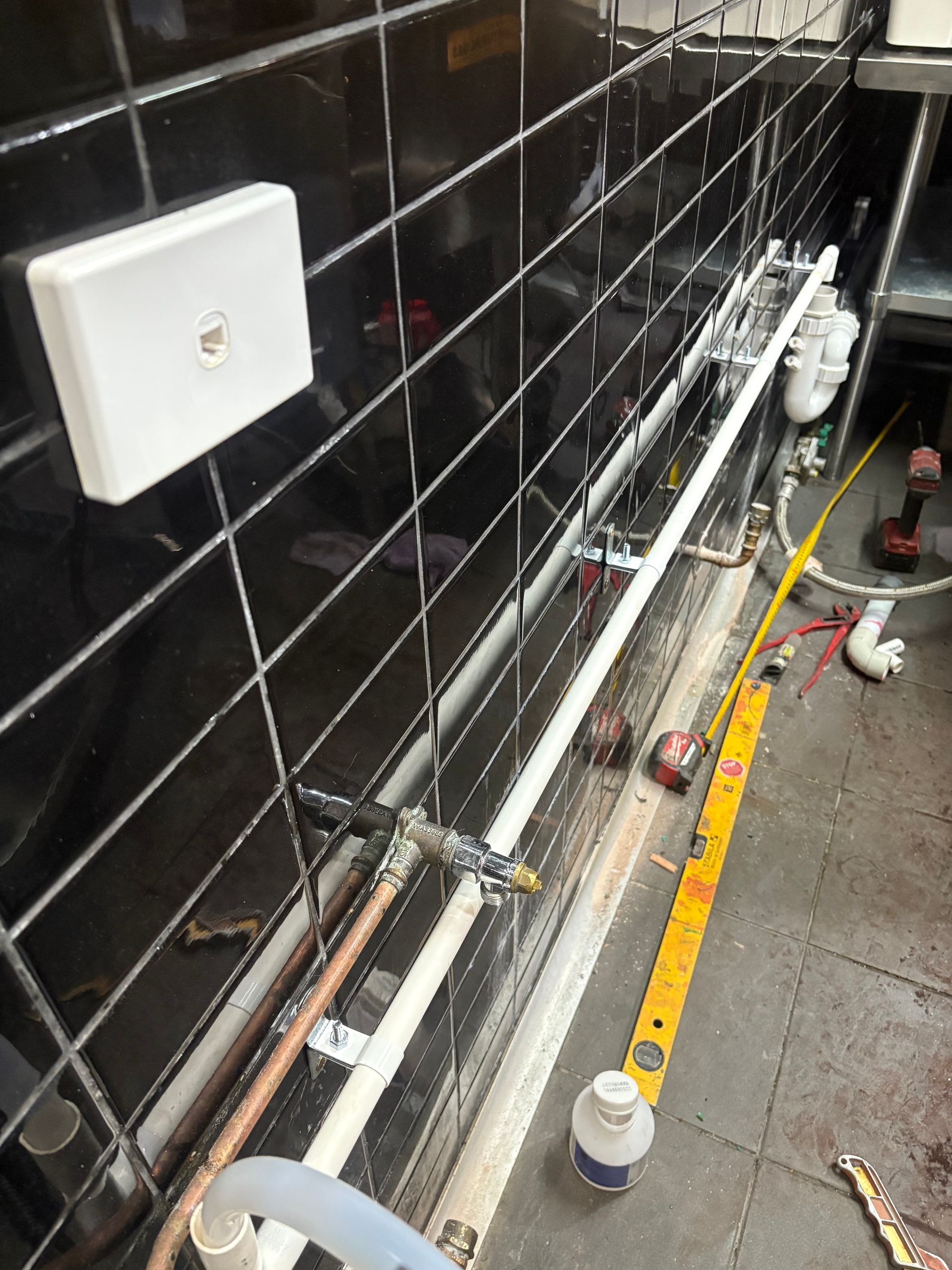 Utility pipes and valves running along a black tiled wall on a rough floor, with a white wall outlet above.