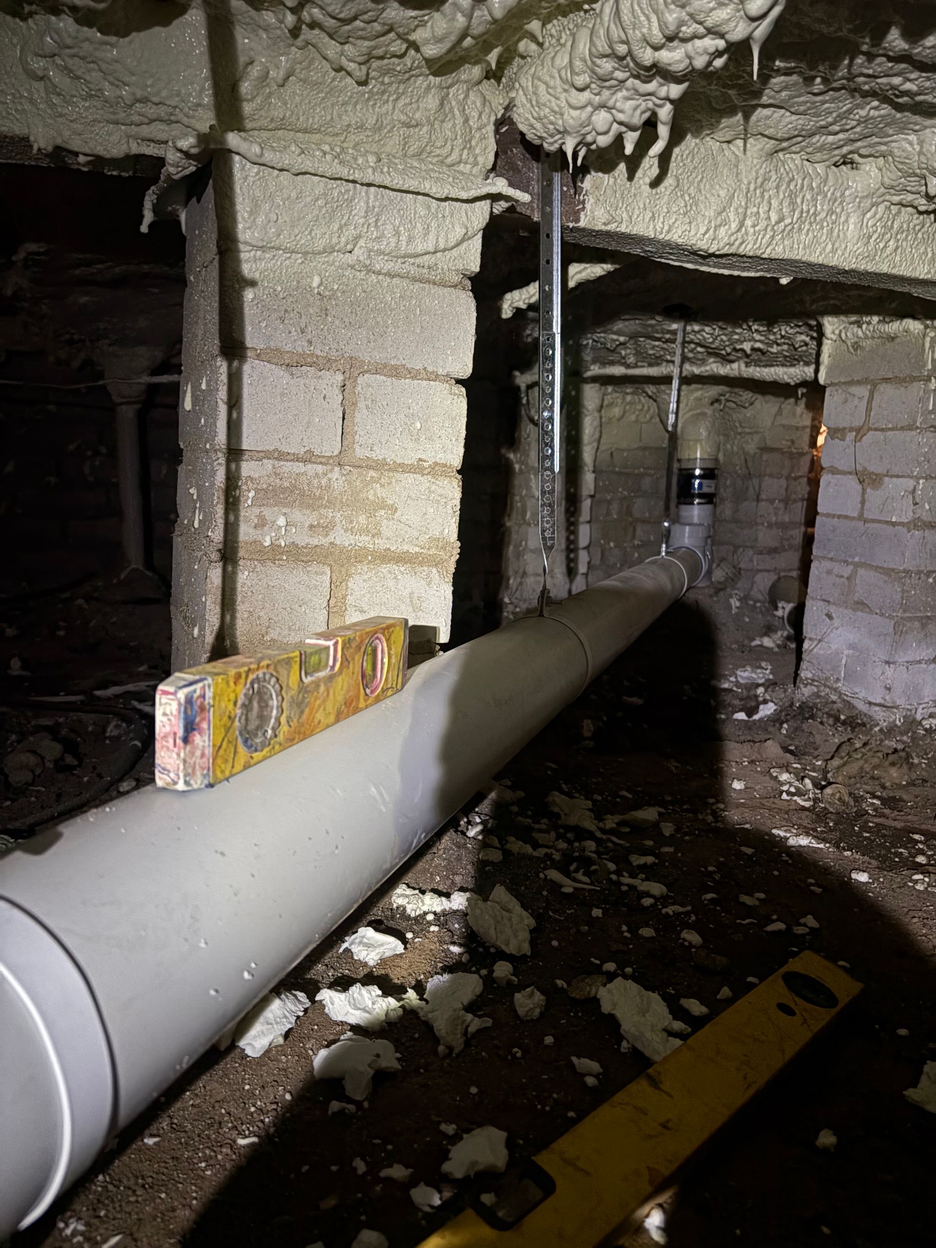 Crawl space with large gray pipe, exposed insulation, and debris on the floor under a low ceiling