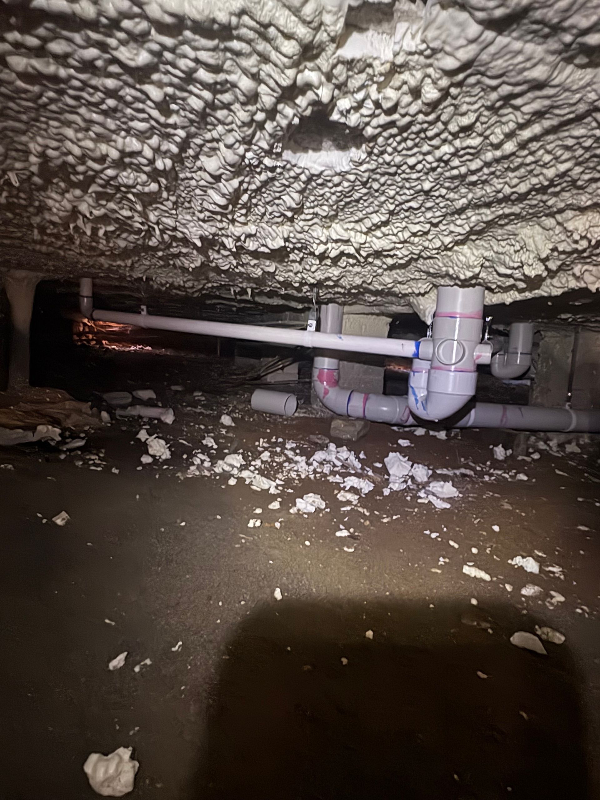 Pipe and plumbing under a low, rough ceiling in a crawlspace, with debris on the ground.