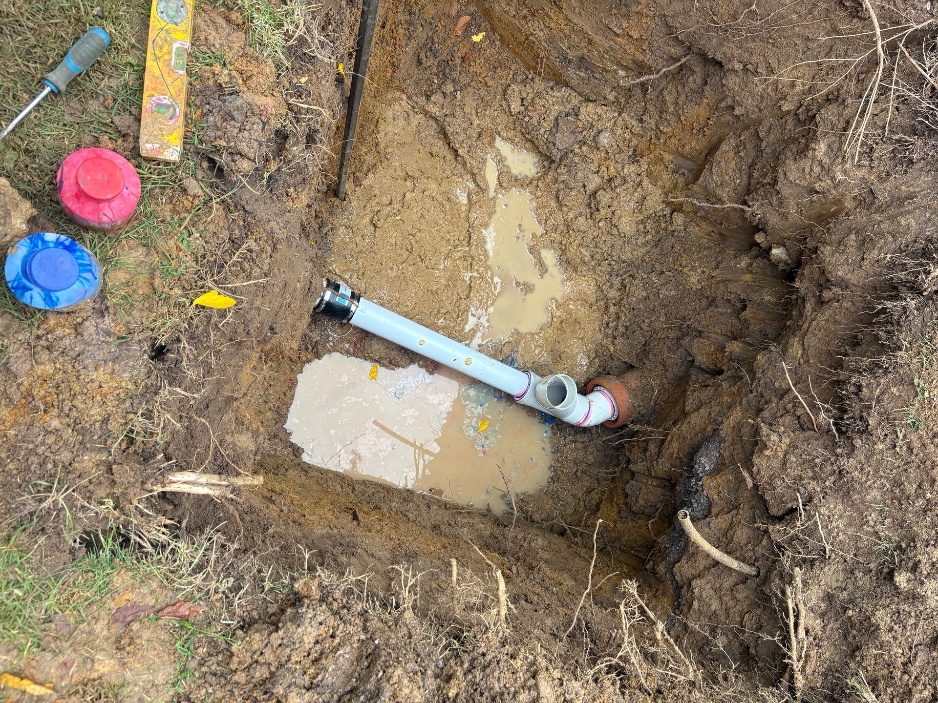 Excavation hole with muddy water and a white PVC pipe; colorful caps and tools lie on the dirt nearby.
