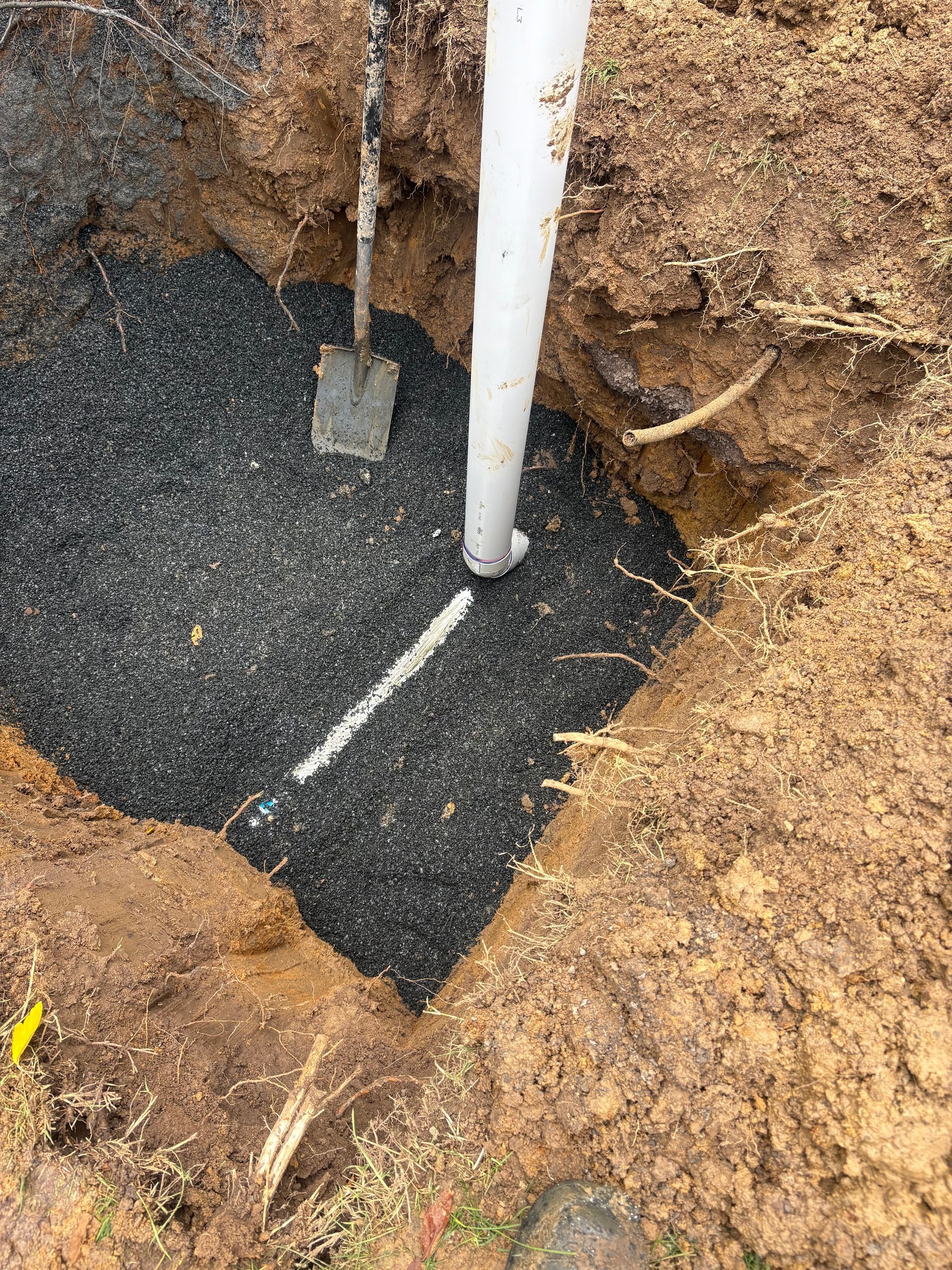 Hole in ground with white PVC pipe and small metal post set in dark gravel fill