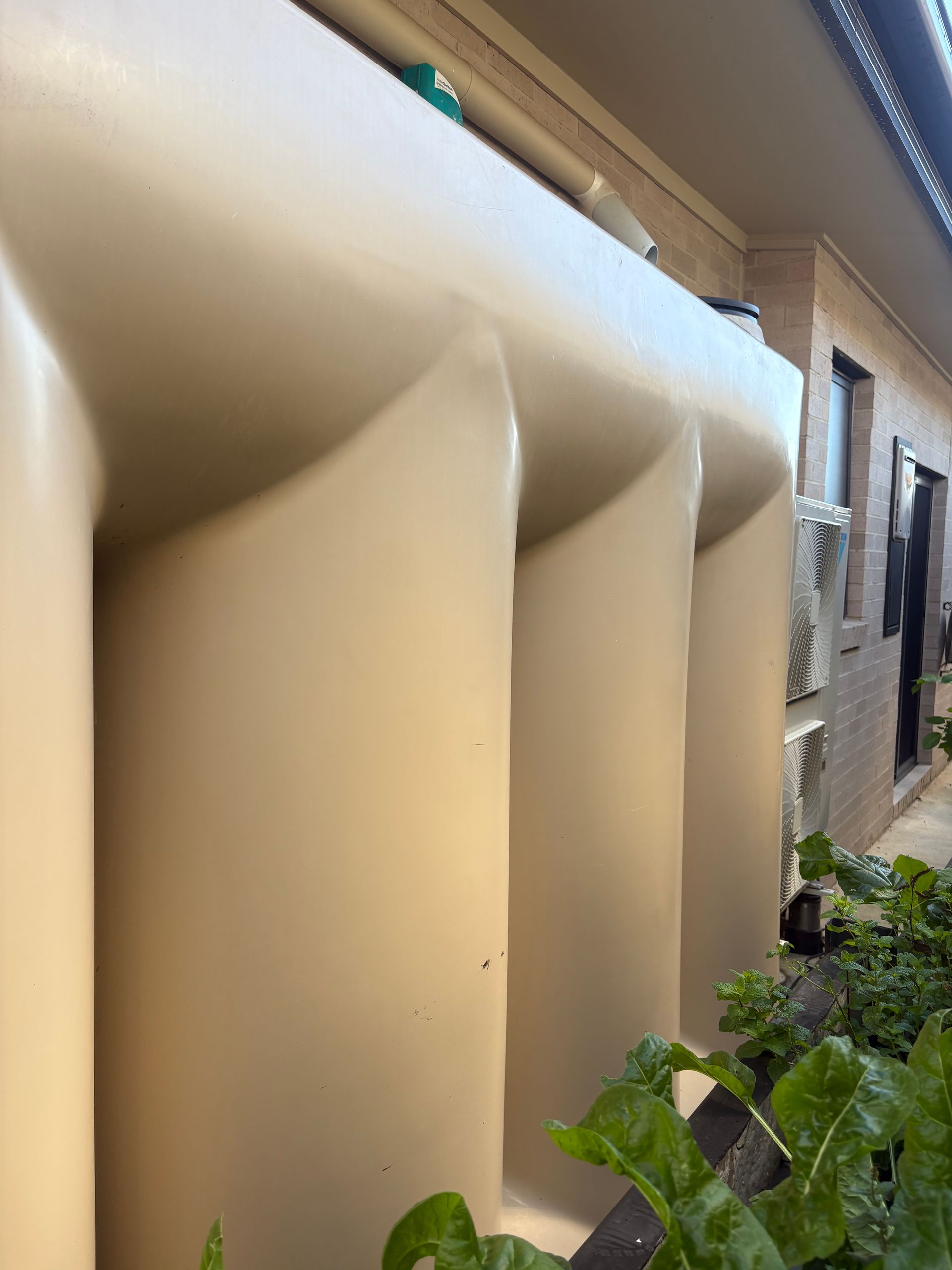 Beige water tanks beside a building, with green plants in the foreground.