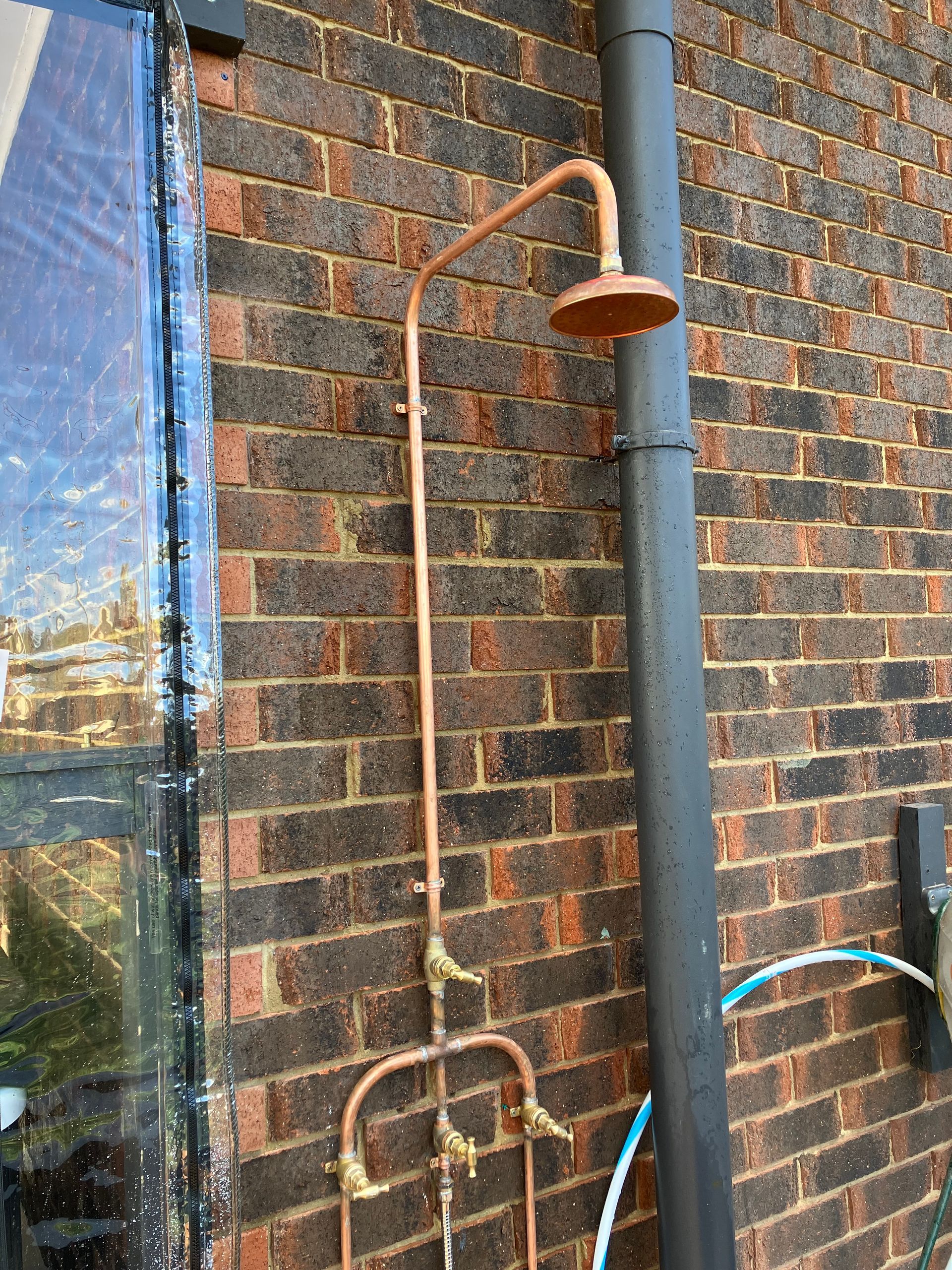 Outdoor shower attached to a brick wall, with copper pipes and a showerhead рядом a window reflection