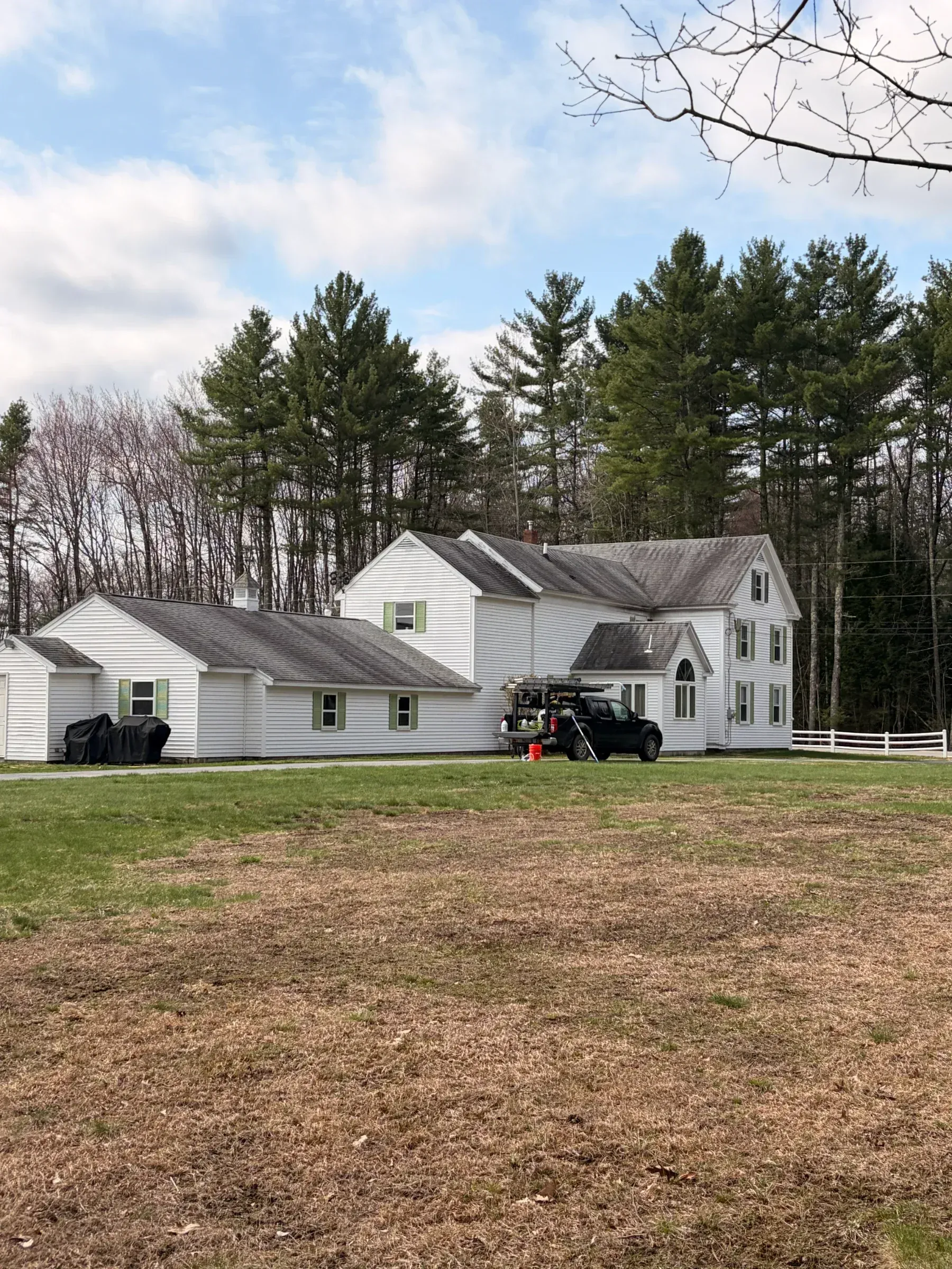 White farmhouse with attached garage and trees on a grassy field under a cloudy sky, with a tractor in front.