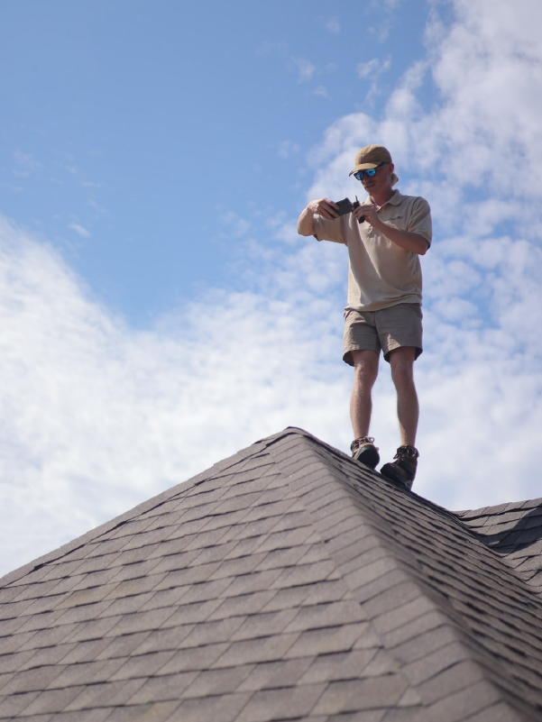 Person standing on a shingled roof, looking at a device against a blue sky with clouds.