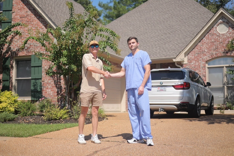 Two men shaking hands in a driveway in front of a brick house and parked SUV