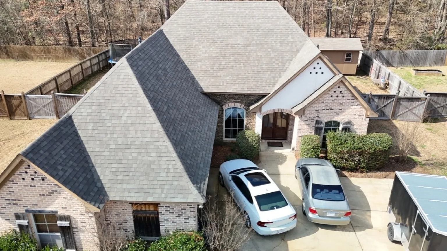 Aerial view of a suburban brick house with two cars parked in the driveway and a fenced yard.