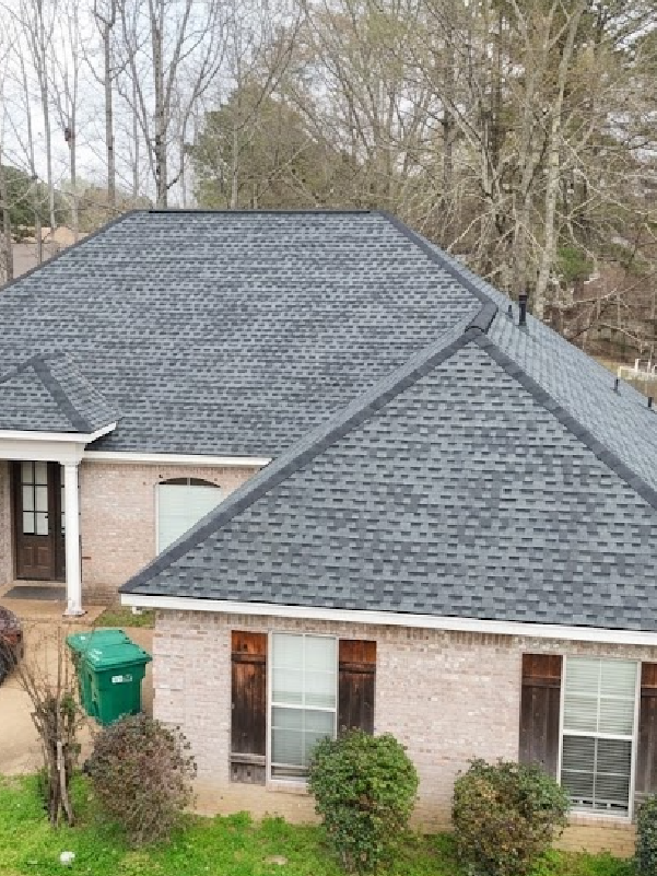 Brick house with a dark gray shingled roof and a green bin in the front yard.