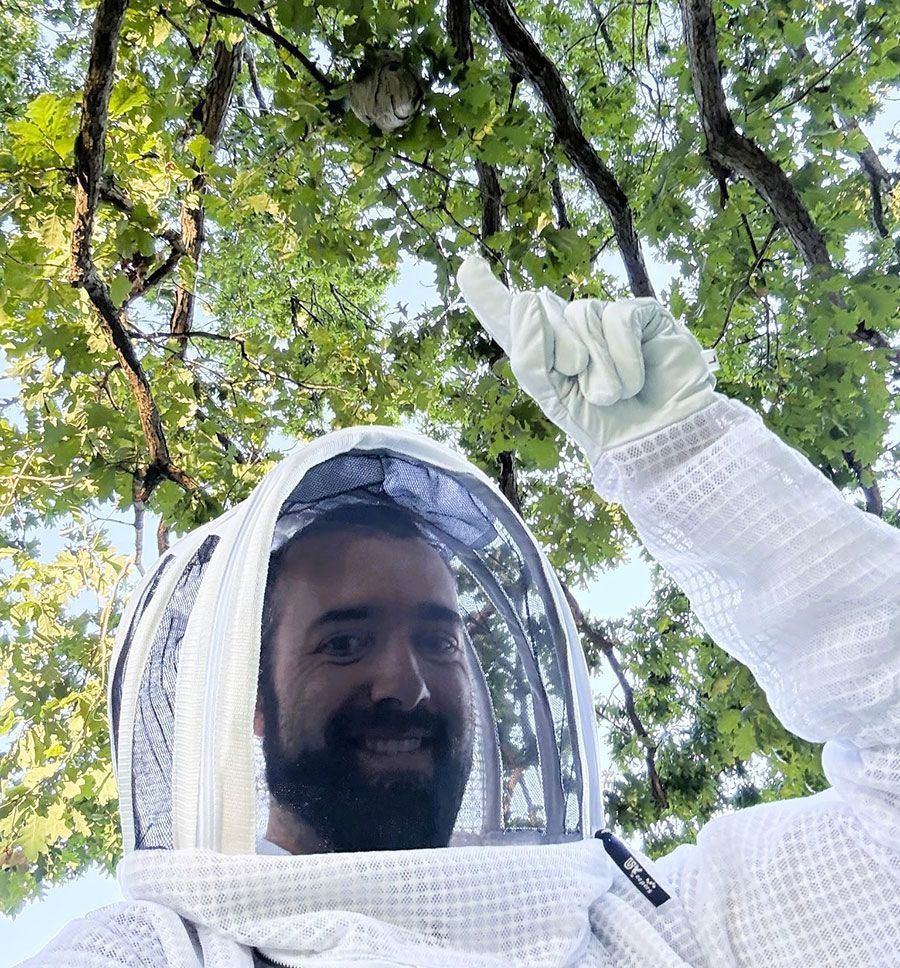 Person in a beekeeping suit and veil smiling and pointing up under leafy tree branches