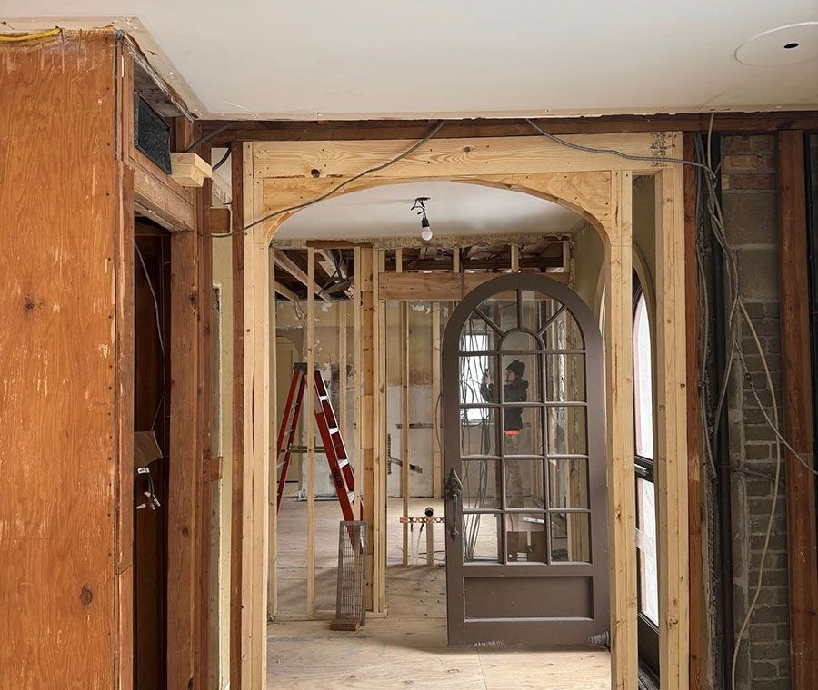 Interior under renovation with exposed framing, arches, a red ladder, and a glass door in the background