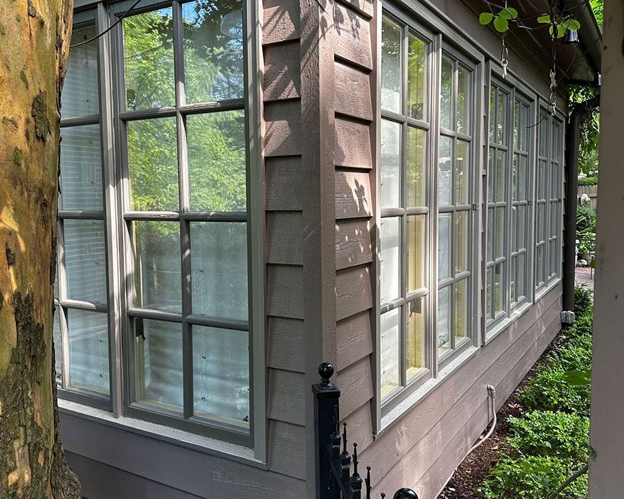 Corner of a house with tall white-paned windows and wood siding beside green plants