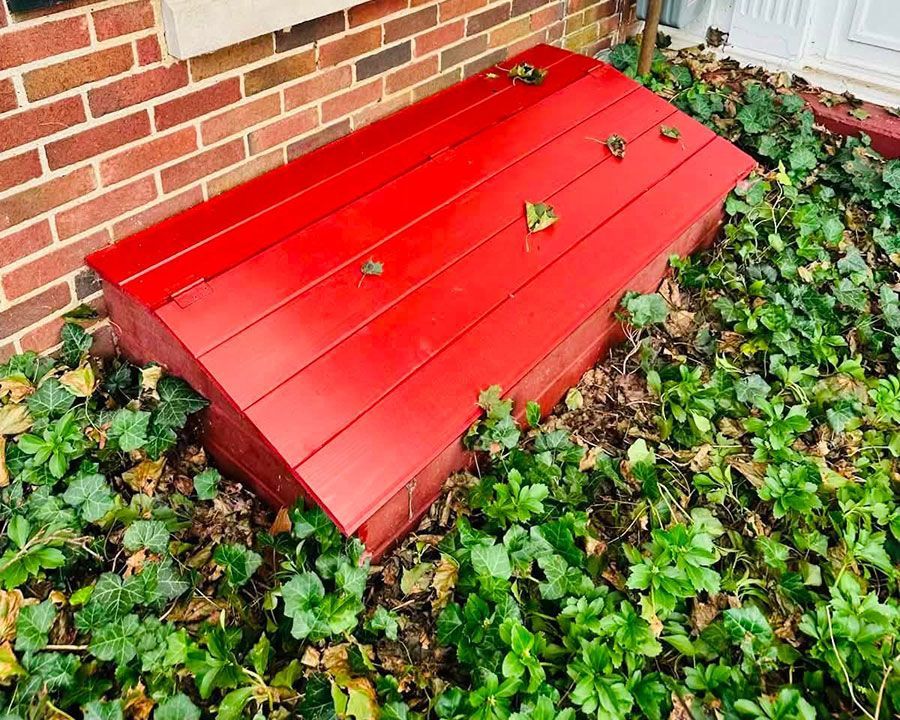 Red wooden planter box against a brick wall, surrounded by green ground cover.