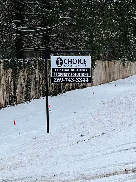 Snowy roadside sign for Choice Custom Builders, with phone number, beside a wooded area.