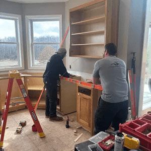 Two people installing a built-in wooden cabinet in a bright room with windows and tools on the floor