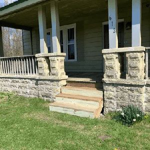 Stone-front porch with steps and white columns on a green house, viewed from the lawn