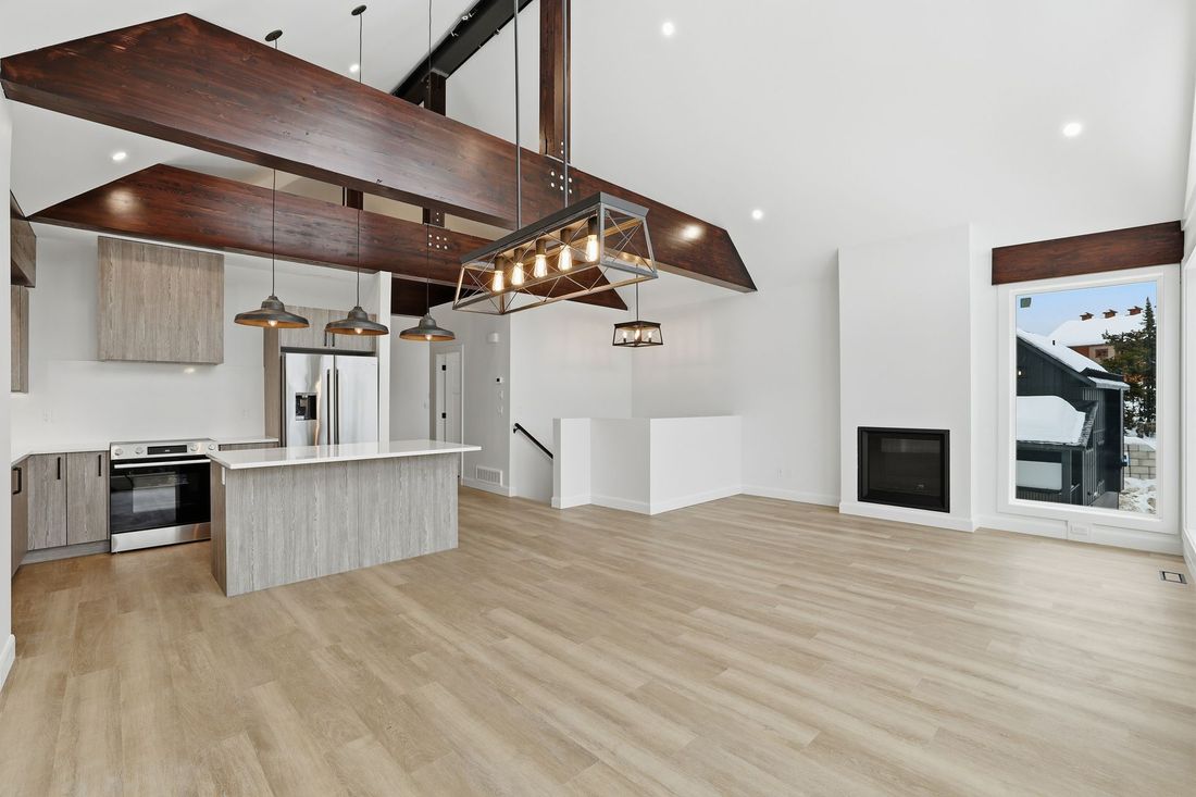 Spacious modern living room with kitchen island, wood beams, light floors, and fireplace.