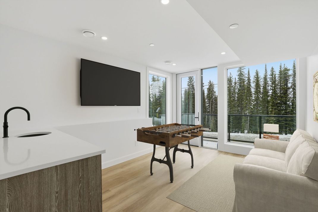 Modern bright living room with wall-mounted TV, foosball table, beige sofa, and large windows overlooking trees