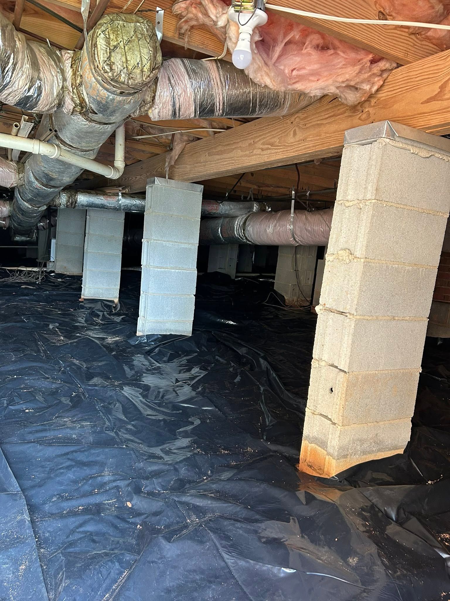 Crawlspace with black vapor barrier on floor, white support posts, exposed insulation and ductwork overhead