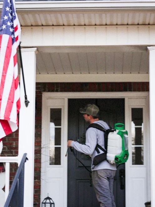 Person with backpack ringing a front doorbell beneath a porch, with an American flag at left.