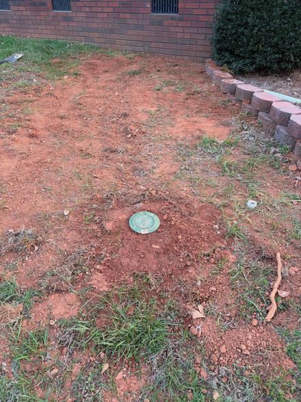 Bare yard with reddish dirt, patchy grass, a green drain cover, and a brick wall edge.