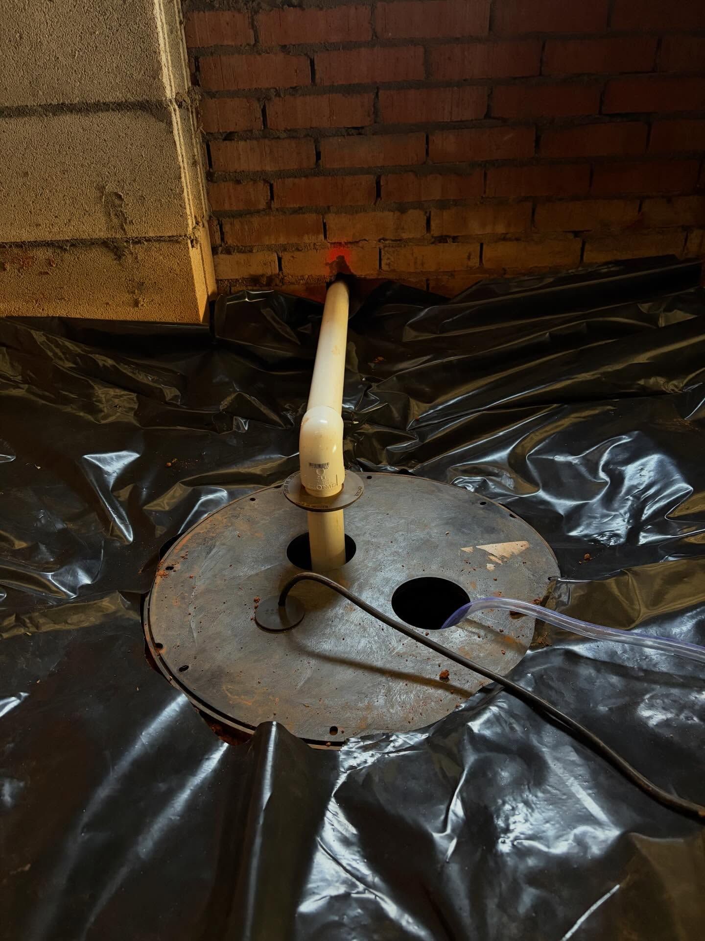 Plumbing pipe emerging from a floor opening covered with black plastic sheeting near a brick wall.