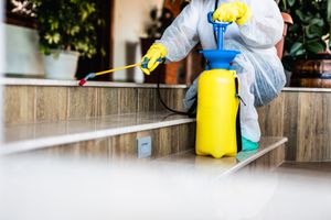 Worker in protective gear spraying disinfectant on outdoor steps with a yellow tank sprayer