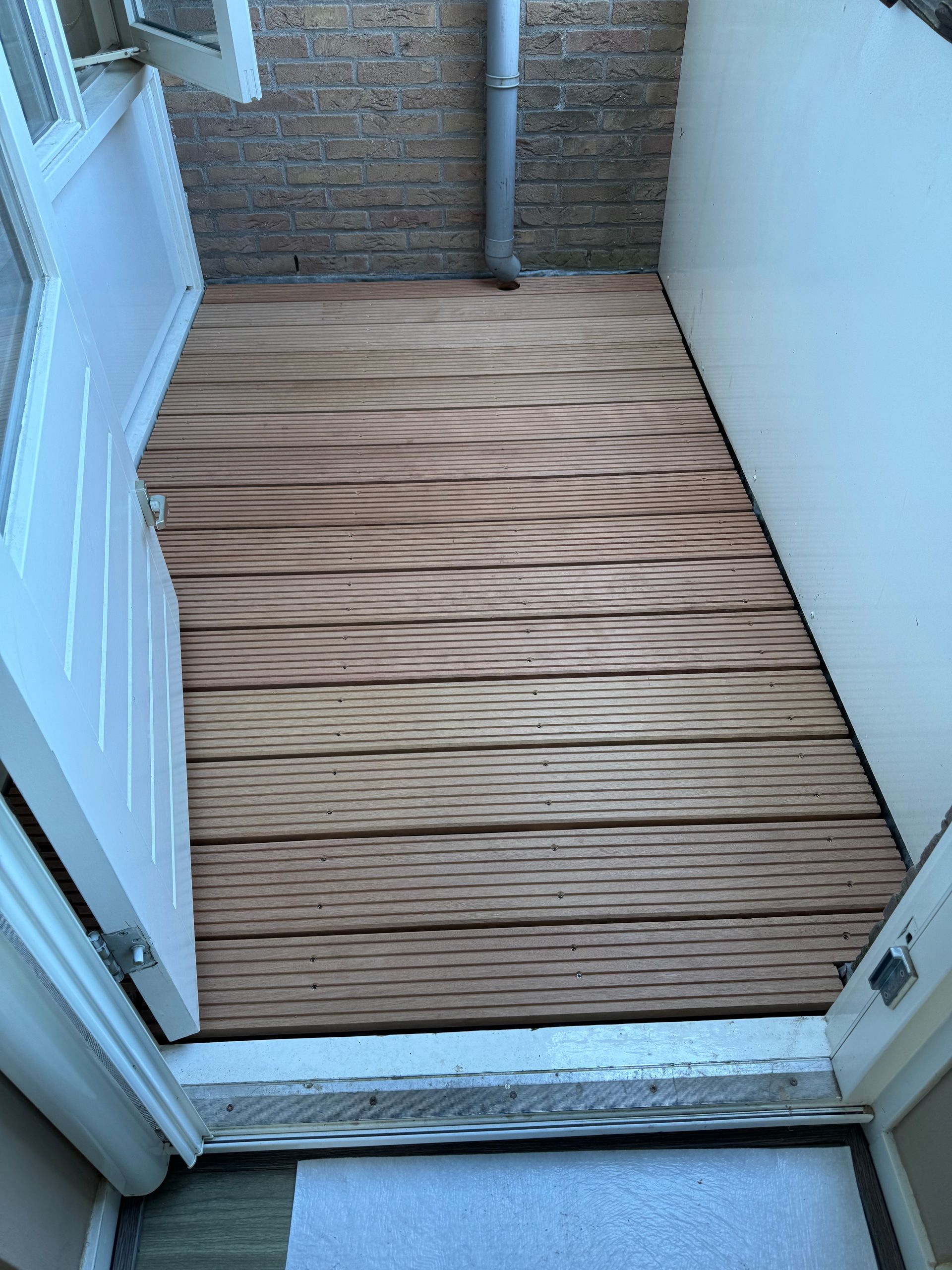 Een open deur geeft toegang tot een klein terras met een houten vlonder en witte muren, dat overgaat in een bakstenen muur met een regenpijp.