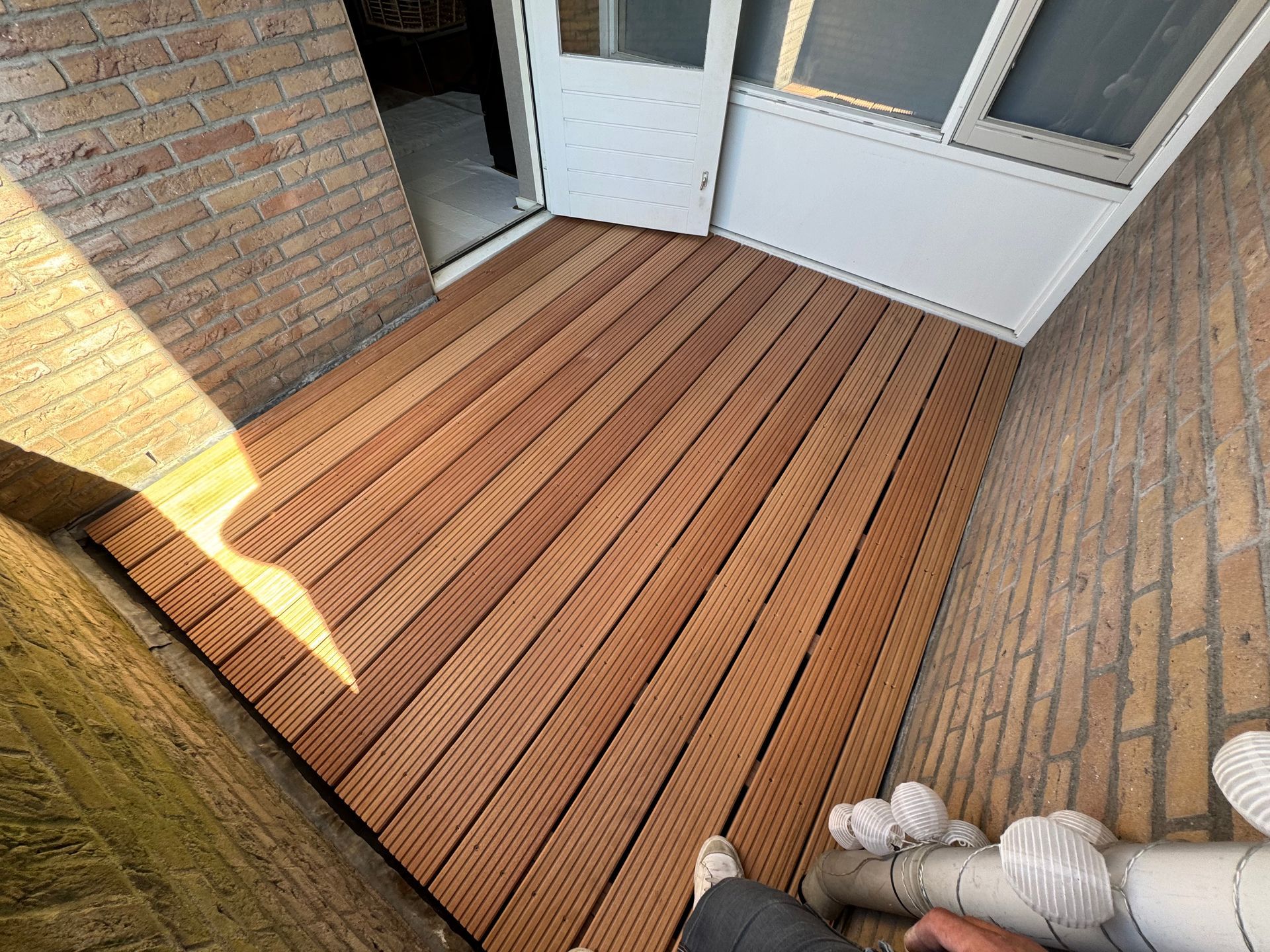 Smal bakstenen terras met houten vlonder, witte deuren en zonlicht dat schaduwen werpt.