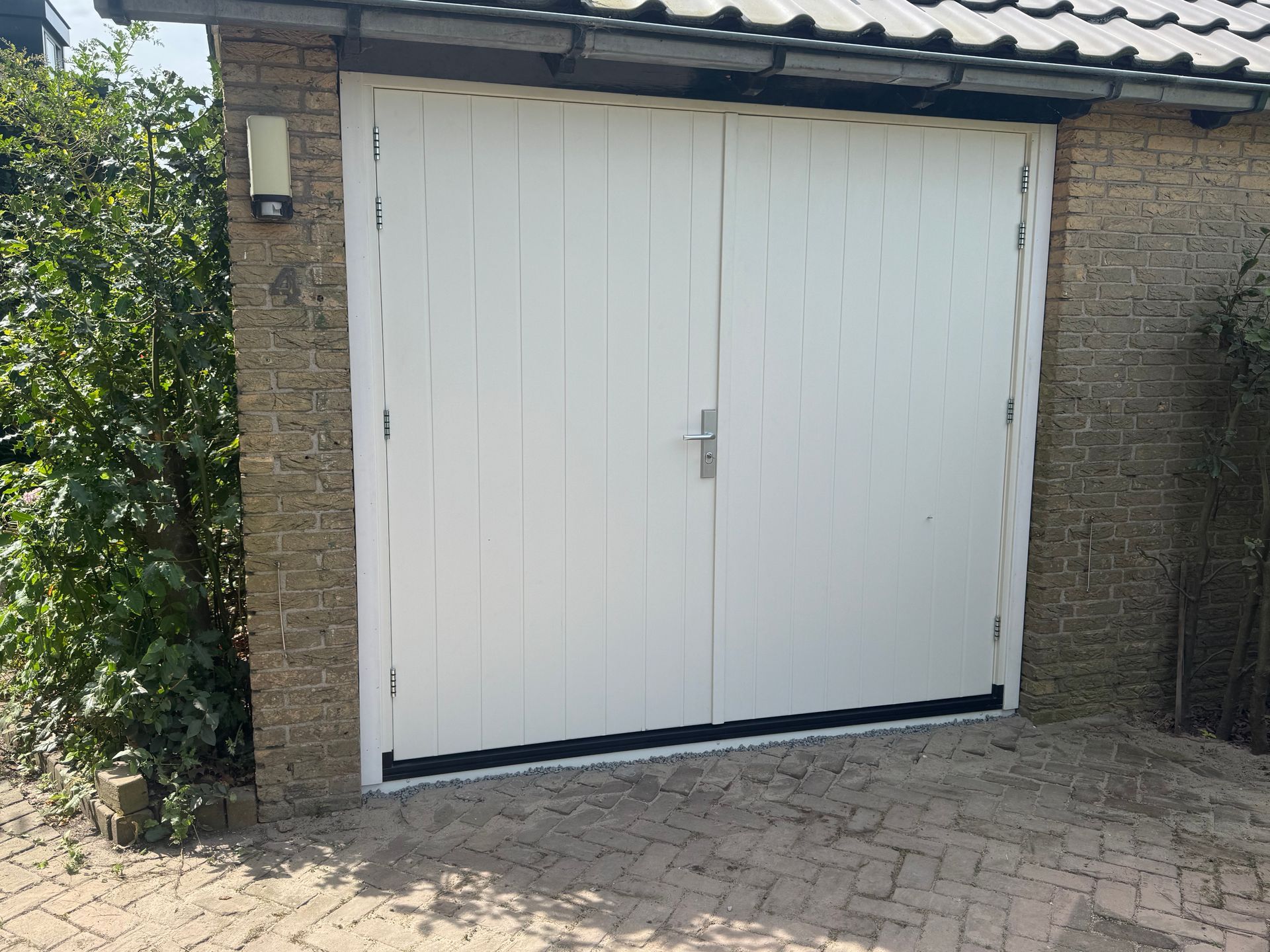 Dubbele witte garagedeuren ingebouwd in een bakstenen muur, met een geplaveide oprit ervoor.