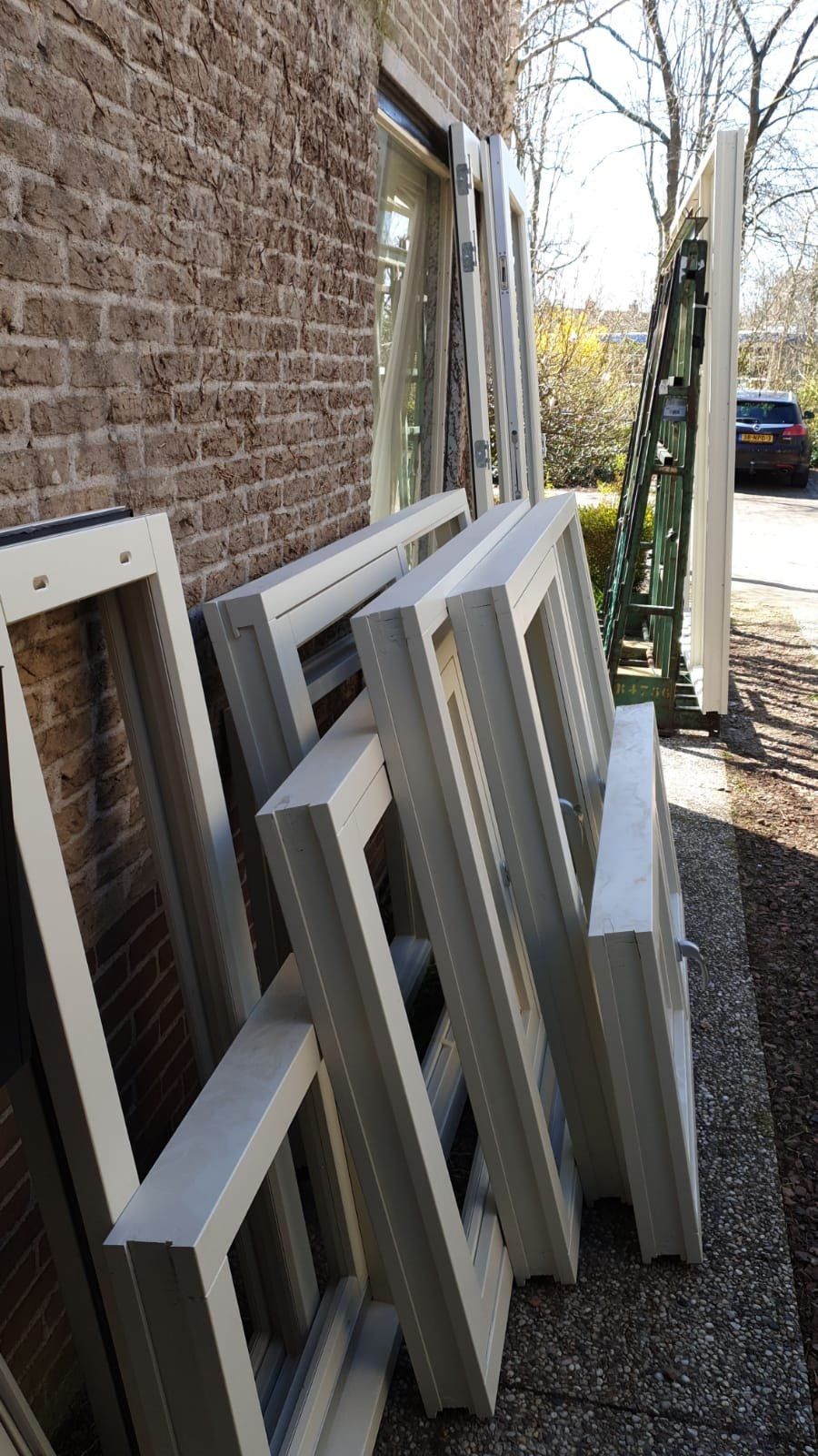 Verschillende ramen met witte kozijnen leunen tegen een bakstenen muur buiten op de stoep.