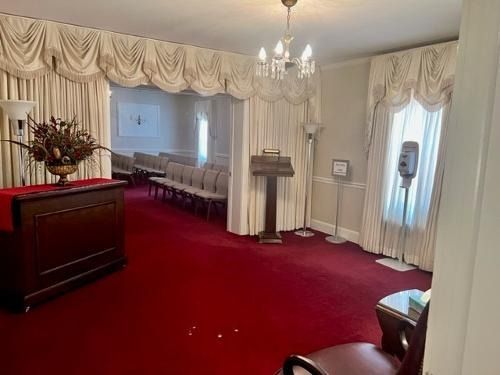 Red-carpeted ceremonial room with white drapes, podium, chandeliers, and rows of chairs along the wall