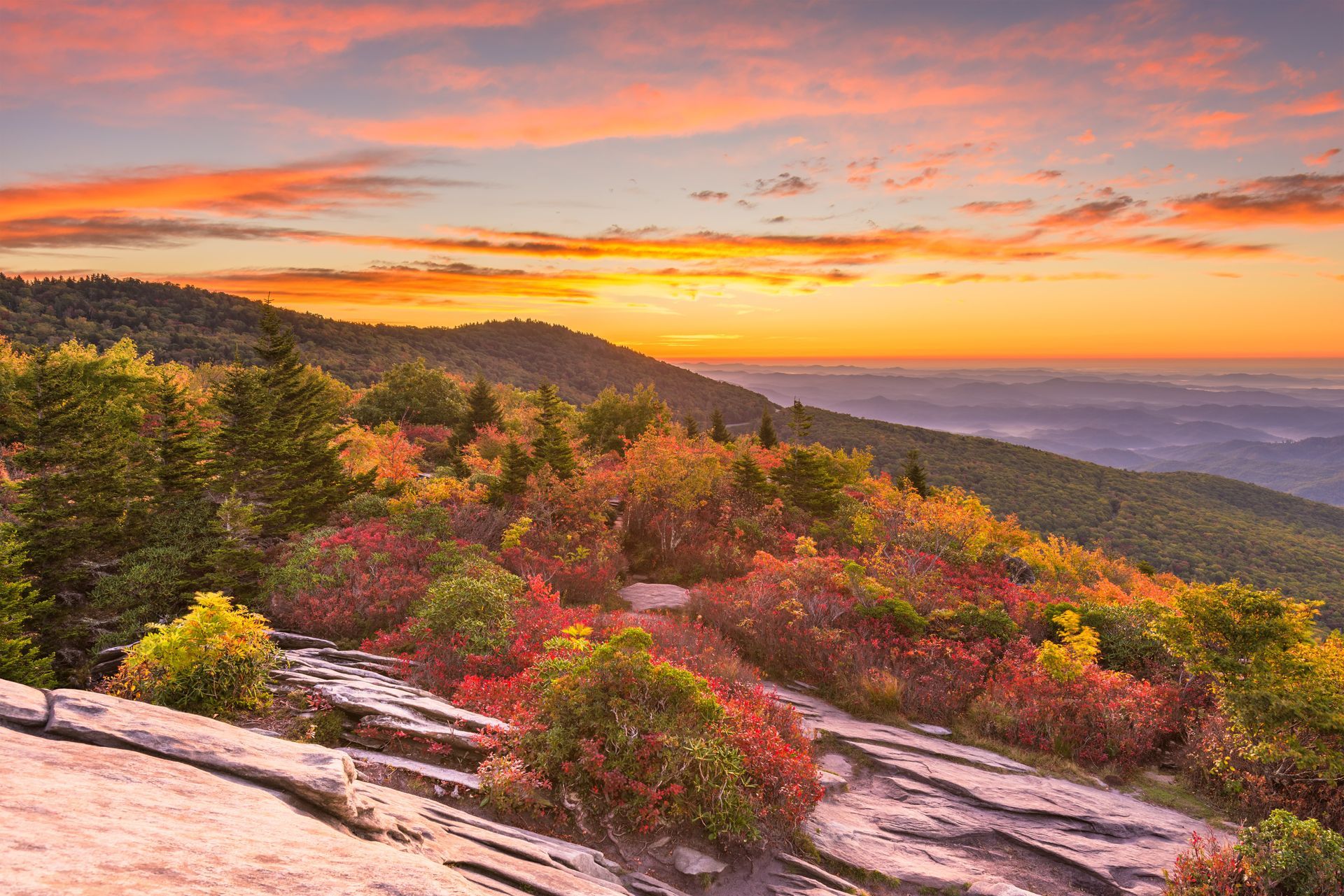 Sunset over rocky hillside with colorful autumn shrubs and distant mountains under a pink-orange sky