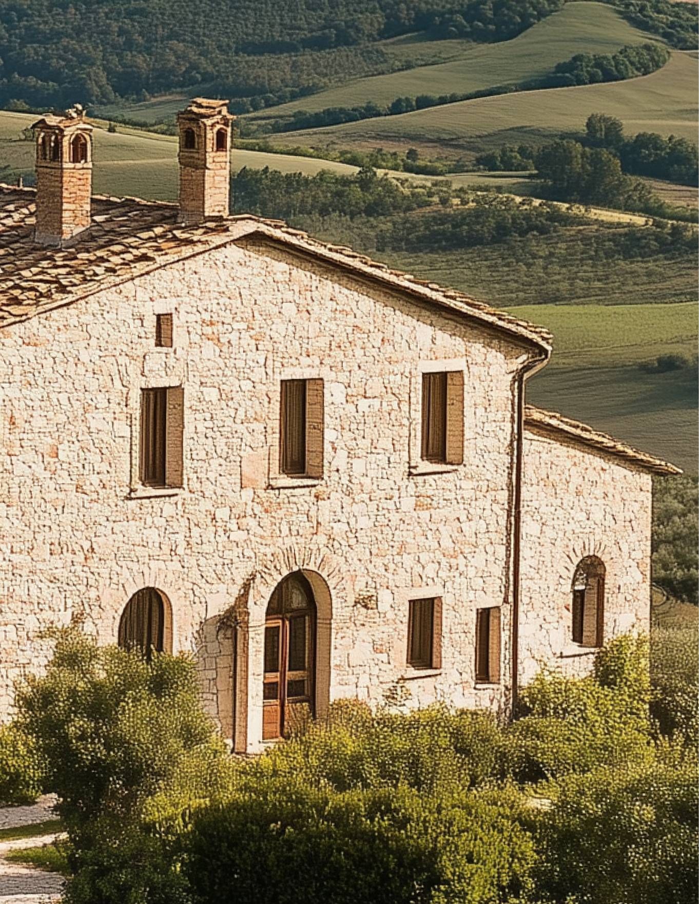 Stone farmhouse with chimneys on a hillside, surrounded by green fields and trees.