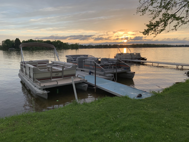 Docked pontoon boats at sunset on a calm lake, with a ramp and grassy shoreline in the foreground.