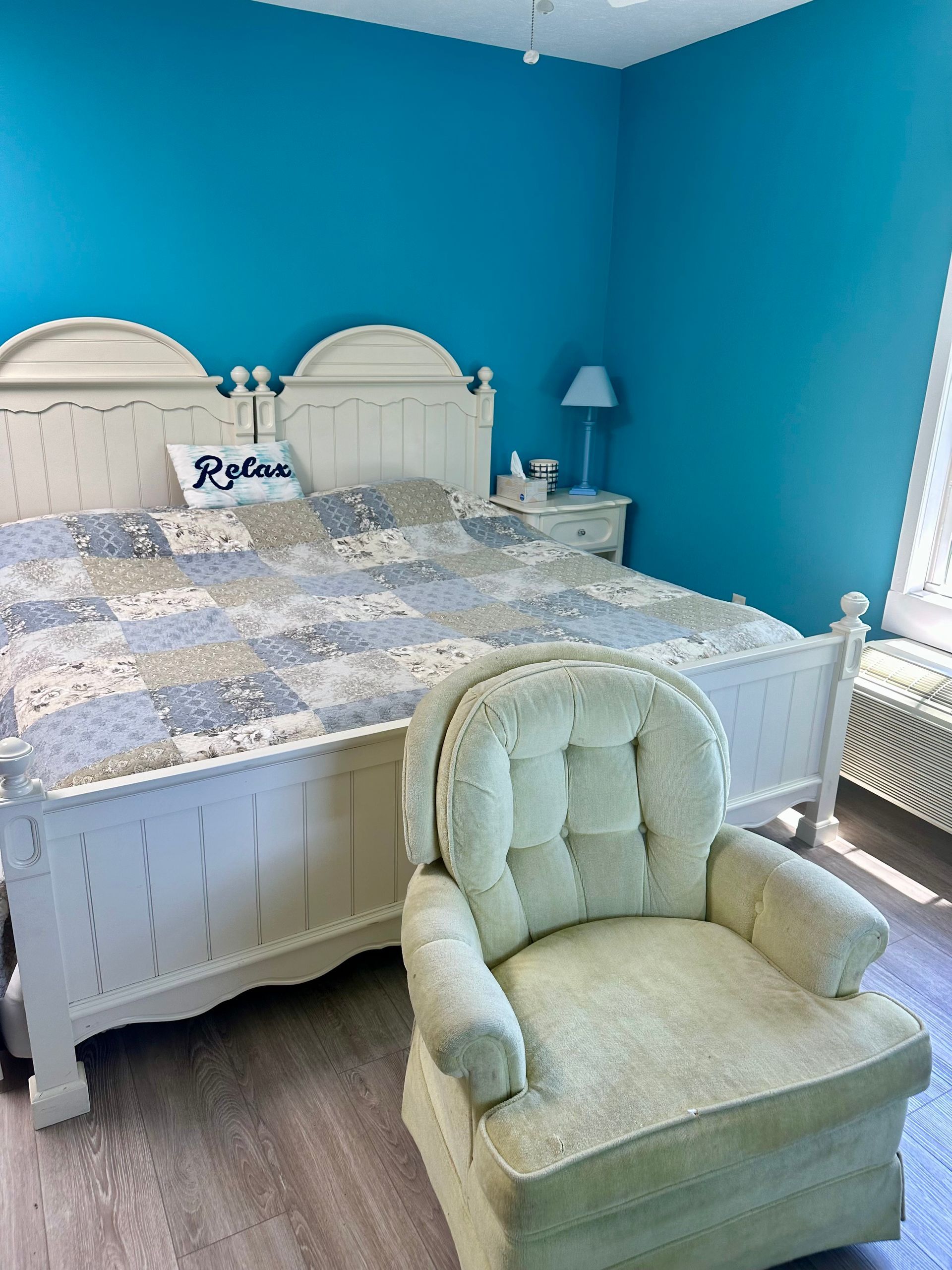 Bright blue bedroom with white bed, patterned quilt, nightstand lamp, and pale green chair in foreground