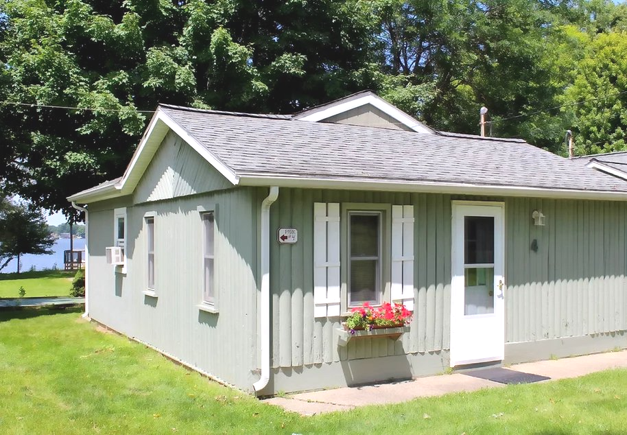 Small light-green cottage with white trim, a white door, and a flower box, beside a grassy yard and trees.