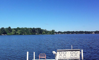 Calm blue lake with a tree-lined shore and a dock with a white bench in the foreground